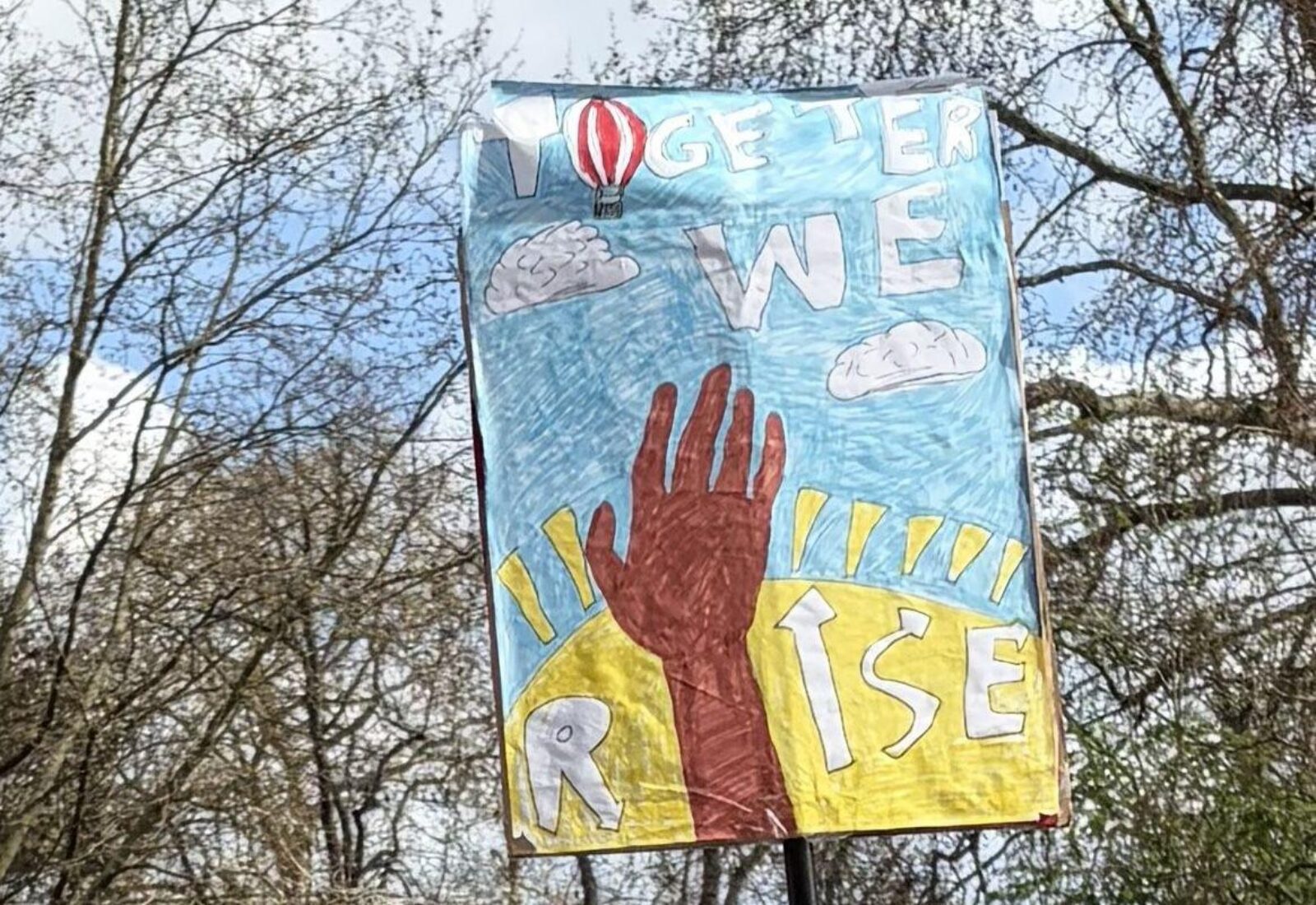 A painted placard at the Together Alliance march, showing a raised hand and a striped hot air balloon with the words TOGETHER WE RISE